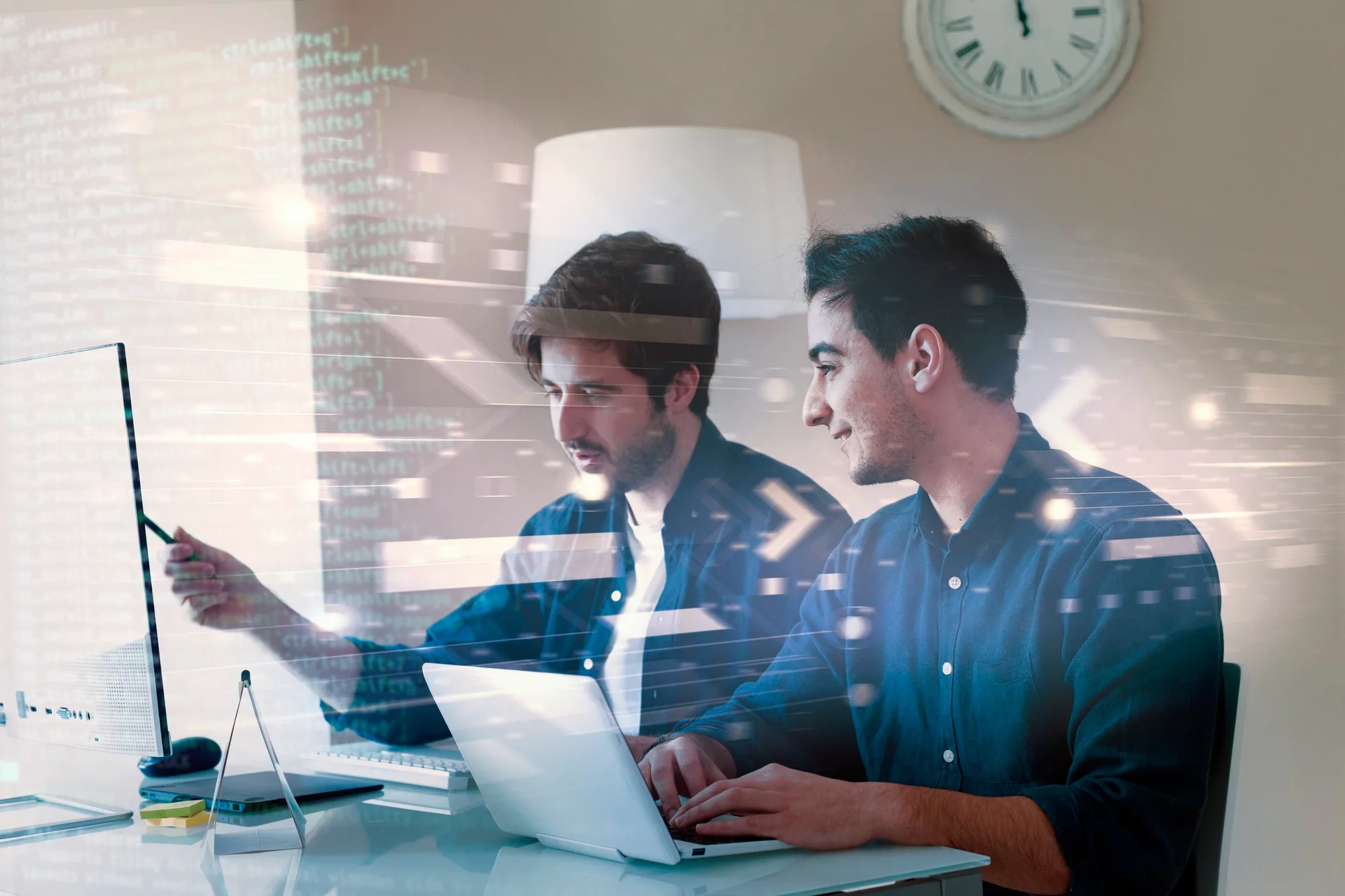 Zwei Personen arbeiten gemeinsam an einem Computer und Laptop mit eingeblendeten Codezeilen.
