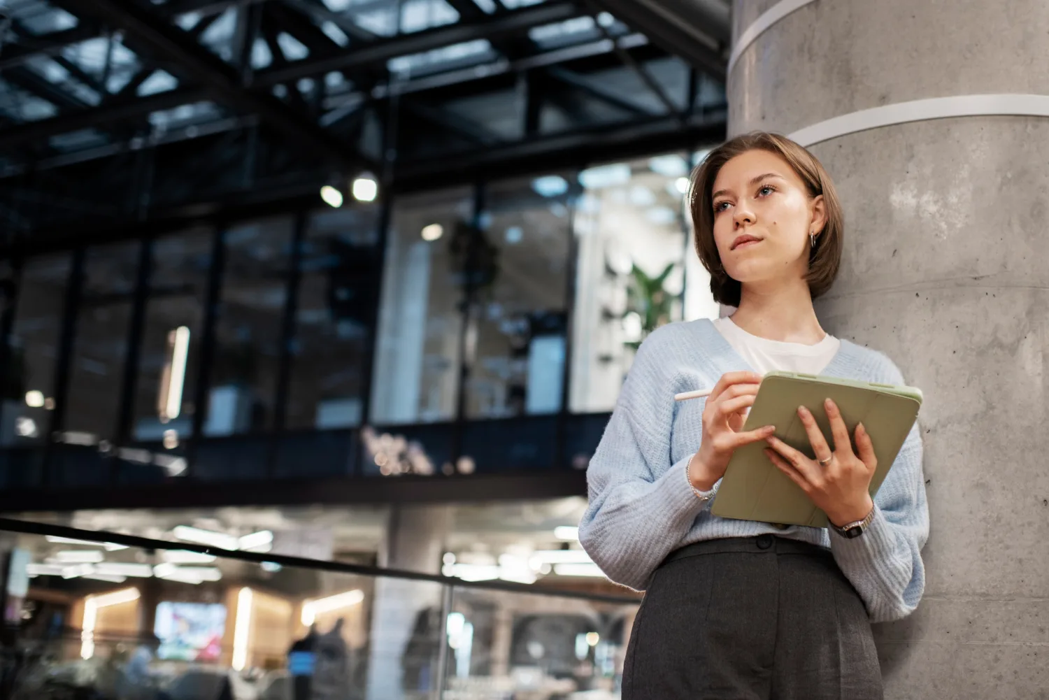 Young woman with a tablet and stylus leaning on a concrete pillar in a modern office, thinking about a product plan