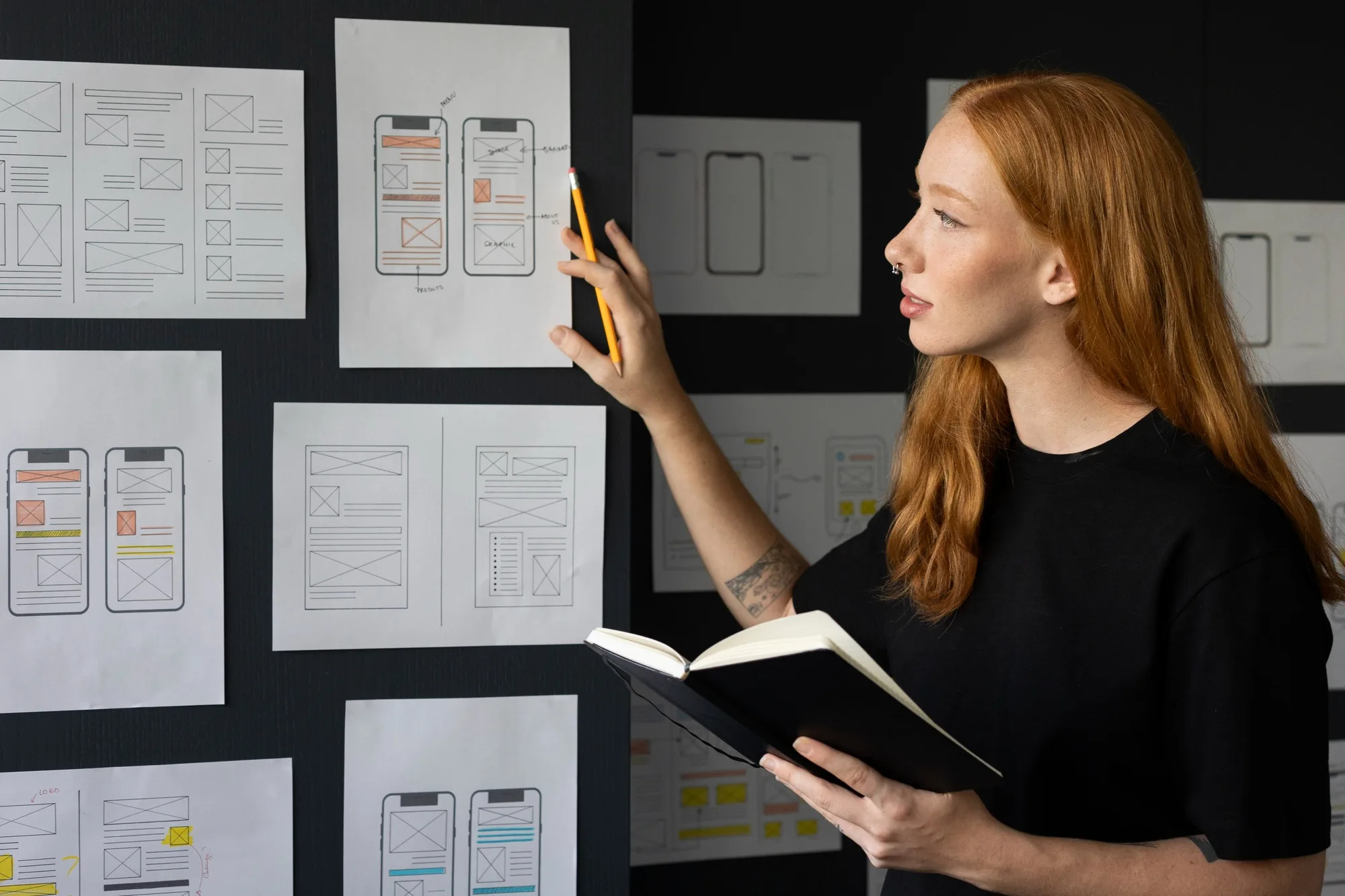 Vrouw wijst naar papieren wireframes van mobiele appschermen aan een muur terwijl ze een notitieboekje vasthoudt