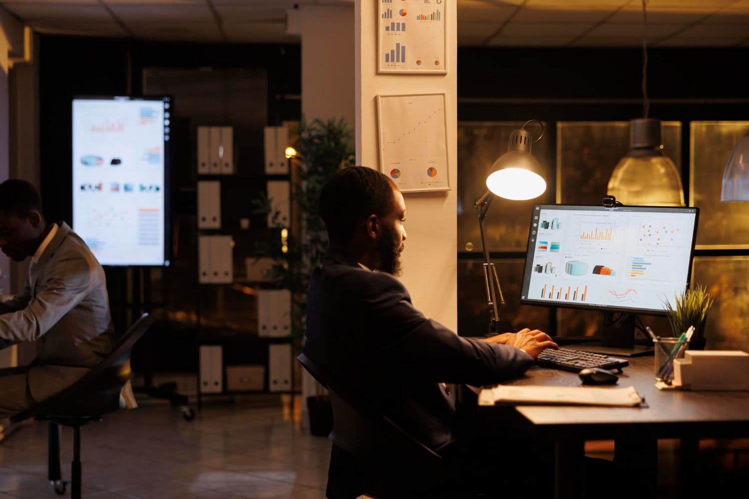 Productanalist die laat doorwerkt aan een bureau voor een beeldscherm en een wandscherm vol grafieken en dashboards