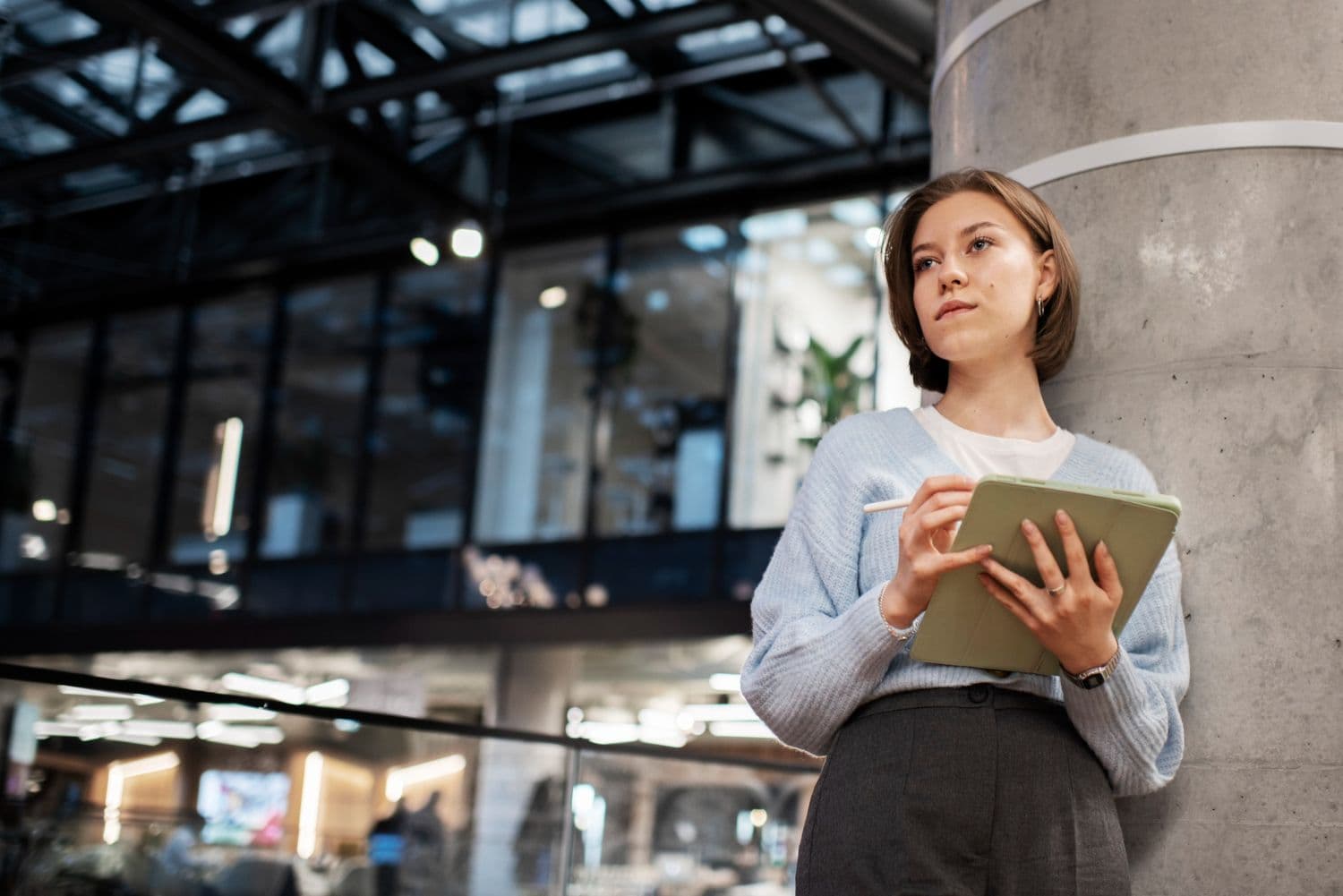 Jonge vrouw met een tablet en stylus die tegen een betonnen pilaar leunt in een modern kantoor en nadenkt over een productplan