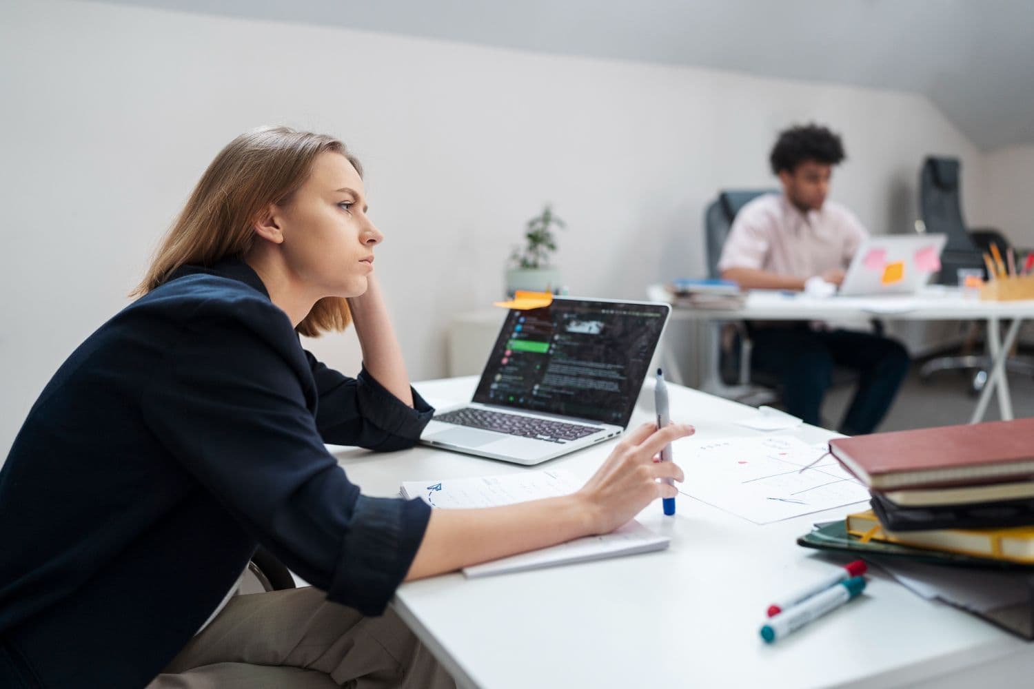 Ontevreden vrouw die haar hoofd op haar hand laat rusten aan een bureau met een laptop, notities en pennen, met een collega achter haar
