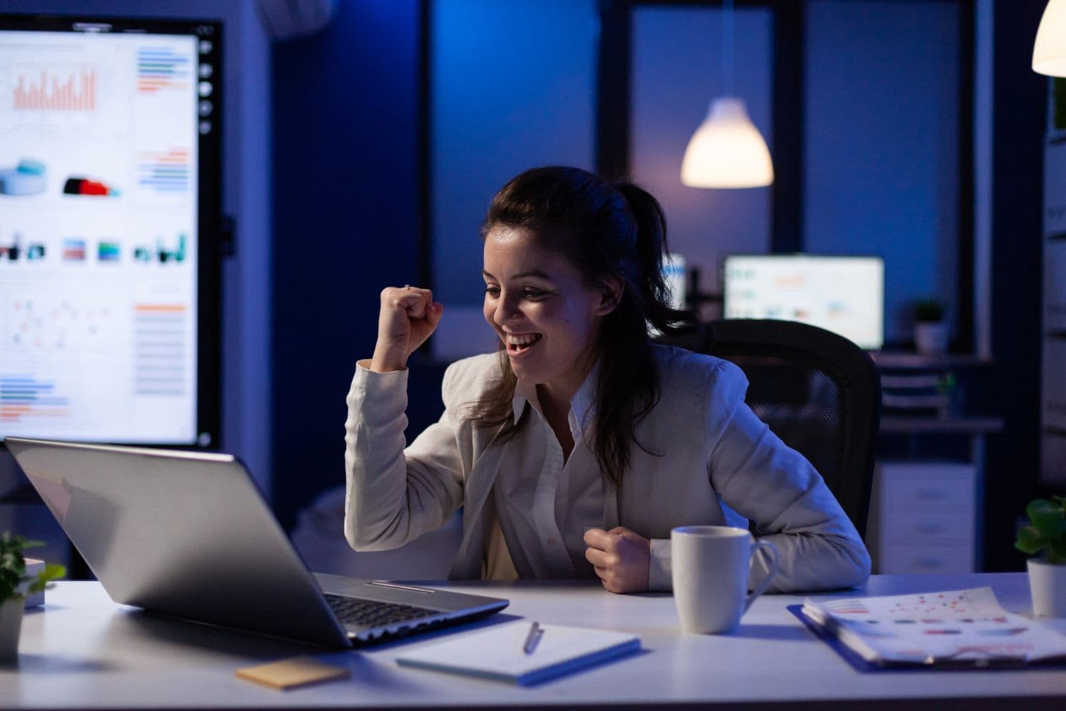 Vrouw die met een opgestoken vuist viert terwijl ze naar haar laptop kijkt, met datagrafieken op een wandscherm achter haar